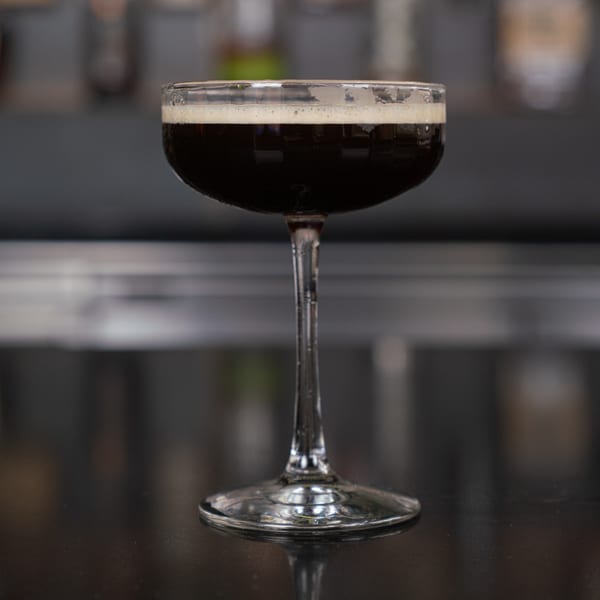A dark cocktail with a foamy top in a coupe glass on a dark, reflective surface.