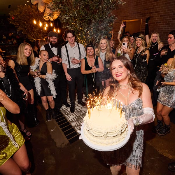 A woman in a flapper dress holds a birthday cake with sparklers, surrounded by smiling guests.