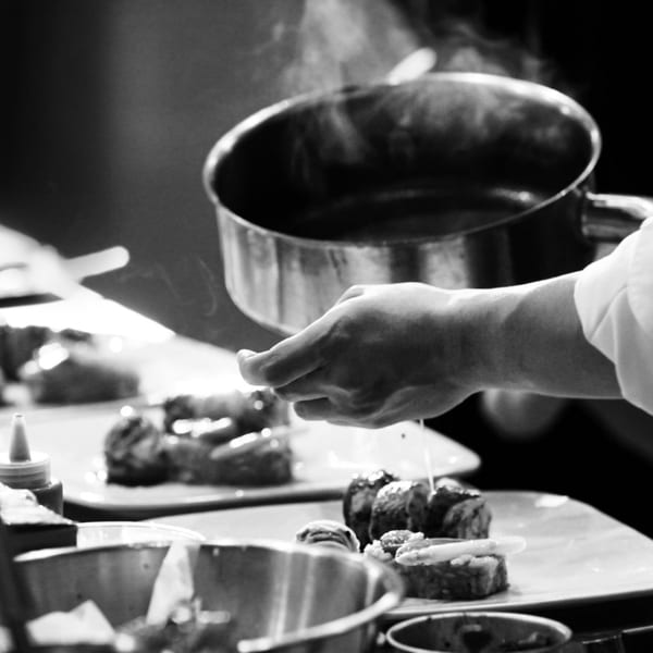Chef's hand pouring sauce from a steaming pot onto American cuisine dishes in a black and white image.