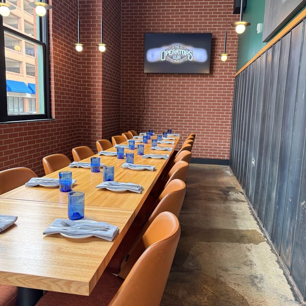 Long wooden dining table set with blue glasses and napkins, brown chairs, brick wall, and TV.