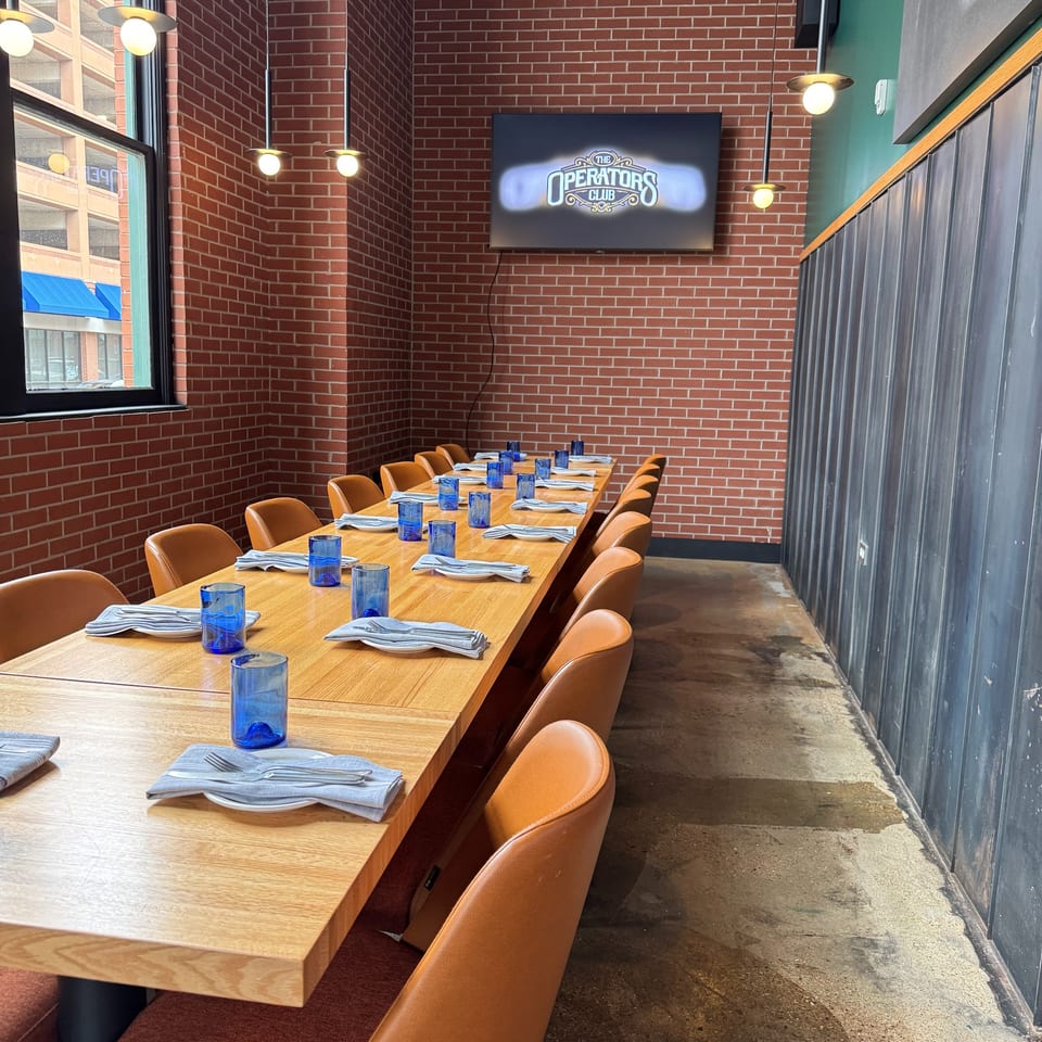 Long wooden dining table set with blue glasses and napkins, brown chairs, brick wall, and TV.