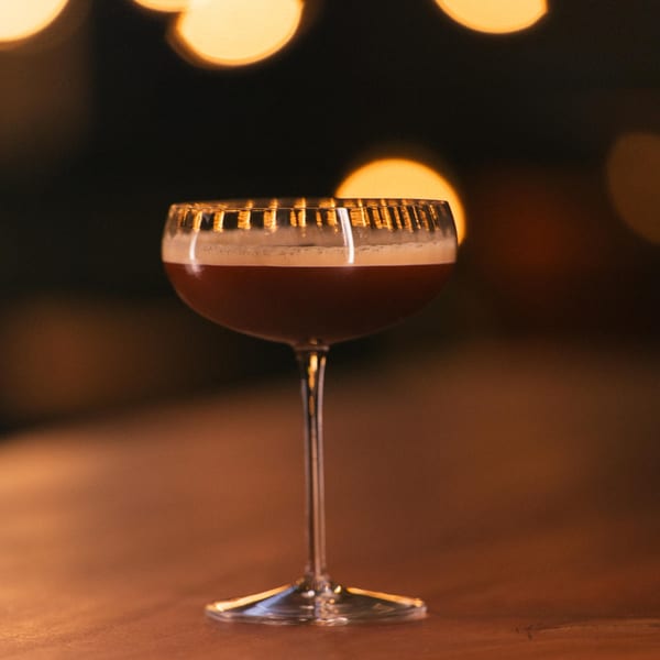 A dark cocktail with a foamy top in a coupe glass, with warm bokeh lights in the background.