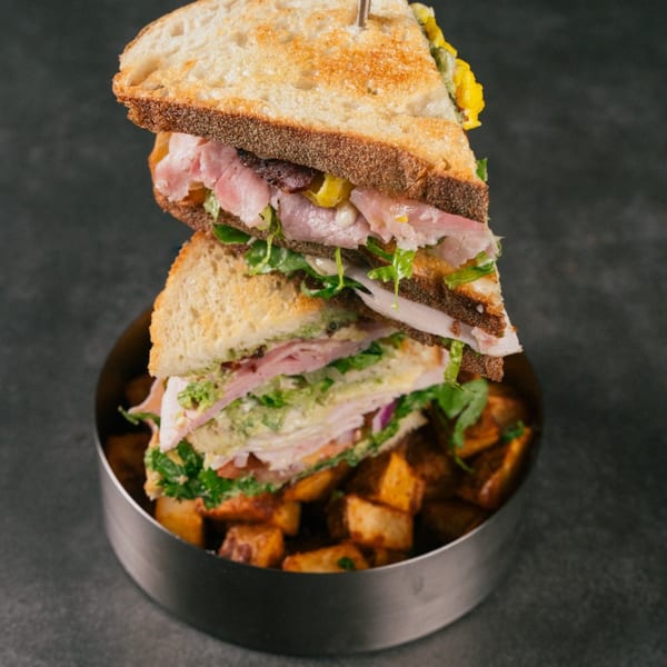 A stacked sandwich with ham, lettuce, and cheese, served with roasted potatoes in a metal bowl.
