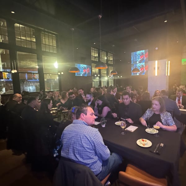 Diners seated at long tables in a dimly lit restaurant with large windows and hanging lights.