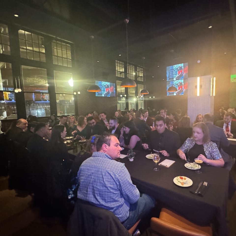 Diners seated at long tables in a dimly lit restaurant with large windows and hanging lights.