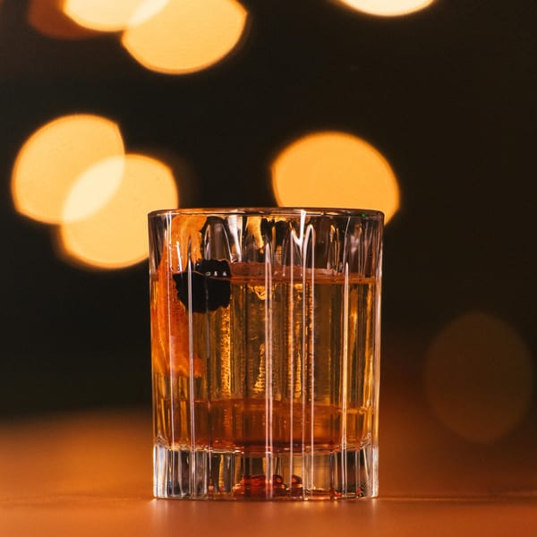 A cocktail with an orange peel and dark berries, against a blurred background of warm lights.