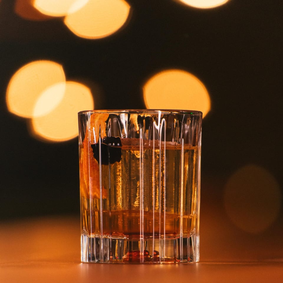 A cocktail with an orange peel and dark berries, against a blurred background of warm lights.
